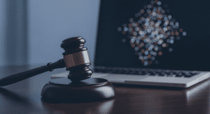 A wooden judge’s gavel rests on a sound block next to an open laptop on a dark table, symbolizing online legal proceedings or digital justice.