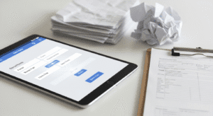 A tablet displaying a form is on a desk next to a clipboard with documents, a stack of papers, and a crumpled paper ball.